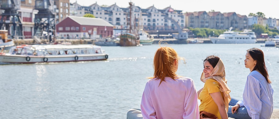 Three female students sit on the edge of a dock, facing the water in the harbour and chatting with each other. It is a sunny day with boats on the water and rows of houses in the background.